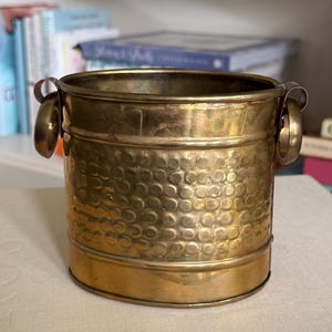 May include: A cylindrical brass bucket featuring a hammered texture on the middle section and two circular side handles. The metal has a polished gold-toned finish with visible horizontal bands near the top and bottom edges of the container.