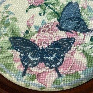 Op de afbeelding: Een ronde, houten lijst met een kruissteek, met twee donkerblauwe vlinders met witte accenten, geplaatst over roze rozen en groene bladeren. De achtergrond is lichtcr&egrave;me met een blauwe rand.