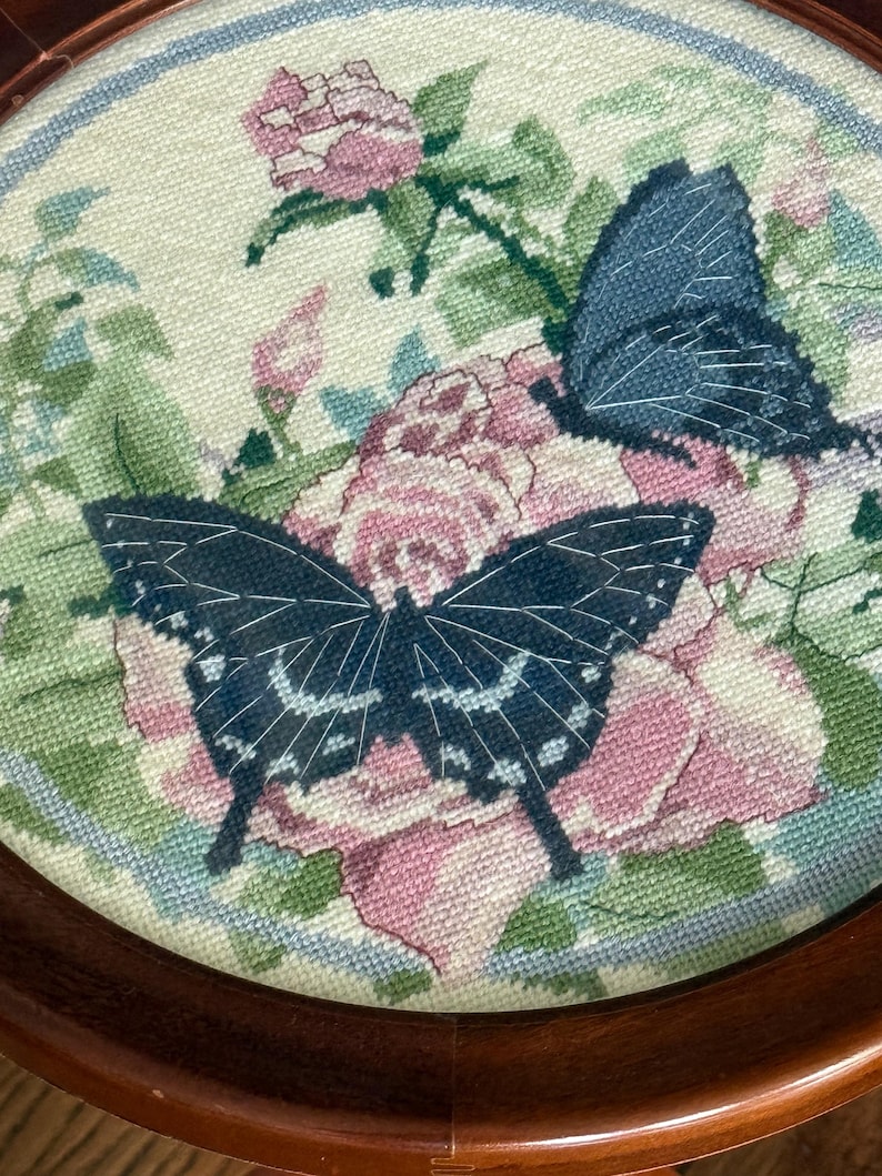 Op de afbeelding: Een ronde, houten lijst met een kruissteek, met twee donkerblauwe vlinders met witte accenten, geplaatst over roze rozen en groene bladeren. De achtergrond is lichtcr&egrave;me met een blauwe rand.