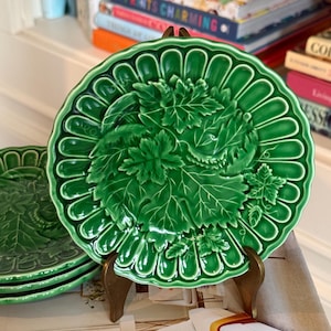 May include: A round, vibrant green ceramic plate featuring a raised, embossed pattern of grape leaves and vines in the center, surrounded by a scalloped, fluted rim. The plate has a glossy finish and is displayed upright on a small stand.