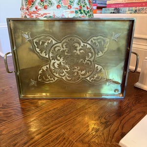 May include: A rectangular brass tray with etched floral and leaf designs. The tray has two handles and a raised edge. The tray is sitting on a wooden floor. Books and a decorative container are in the background.