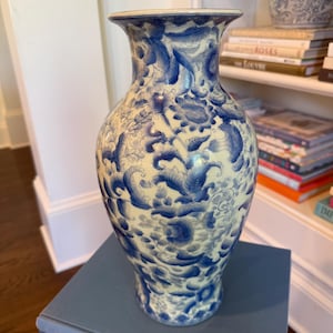 May include: A tall, ceramic vase with a cream-colored base and intricate blue floral patterns. The vase has a wide body that tapers towards the base and a flared rim. The design features stylized flowers and leaves, creating a classic, decorative piece.