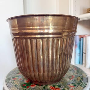 May include: A large, antique brass planter with vertical ribbed detailing. The planter has a wide, rounded shape and a slightly flared rim. It sits on a decorative, round ceramic stool with a floral pattern. The brass has a warm, aged patina.