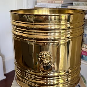 May include: A polished brass cylindrical bucket featuring horizontal ribbed detailing along the top and bottom edges. The front of the vessel is adorned with a decorative lion head emblem holding a circular ring in its mouth, creating a handle.