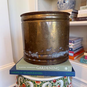 May include: A weathered brass planter sits atop two gardening books and a decorative garden stool. The books are titled "Martha Stewart's Gardening" and "Gardens of the French Riviera." A blue and white ceramic jar is visible on a shelf in the background.