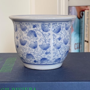 May include: A small, white ceramic planter with a blue floral pattern. The design features stylized blue flowers and vines against a white background. The planter has a rounded shape with a slightly flared rim, suitable for small plants or succulents.