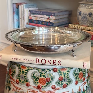 May include: A silver-plated serving dish with a glass insert, resting on a book titled "The Glory of Roses." The dish has a decorative beaded edge and small feet. The background includes a stack of books and a decorative ceramic garden stool.