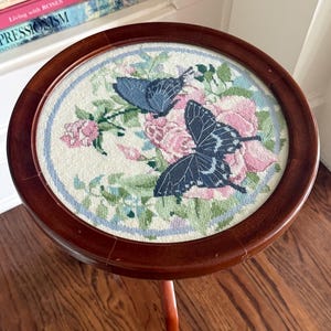 Op de afbeelding: Een kleine, ronde houten bijzettafel met een naaldkunst bovenblad. De naaldkunst toont twee blauwe vlinders te midden van roze rozen en groen gebladerte. De tafel heeft een donkerbruine afwerking en een enkele, gebogen poot.