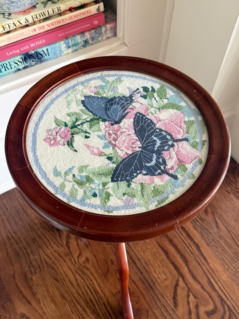 Op de afbeelding: Een kleine, ronde houten bijzettafel met een naaldkunst bovenblad. De naaldkunst toont twee blauwe vlinders te midden van roze rozen en groen gebladerte. De tafel heeft een donkerbruine afwerking en een enkele, gebogen poot.