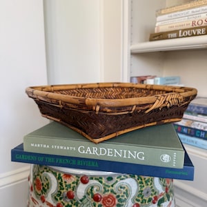 May include: A square, woven bamboo basket with a dark brown finish sits atop a stack of books. The books include "Martha Stewart's Gardening" and "Gardens of the French Riviera." The basket is placed on a decorative ceramic garden stool.