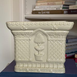 May include: A cream-colored ceramic planter with a rectangular shape and a glossy finish. The exterior features a raised relief design of a potted topiary tree centered within an arched frame, flanked by lattice patterns, and topped with bee motifs.