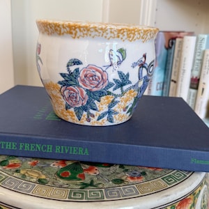 May include: A white ceramic planter with a floral design featuring pink roses, blue leaves, and birds. The rim has a textured, speckled orange-yellow pattern. The planter is sitting on a blue book titled "THE FRENCH RIVIERA".