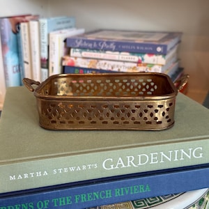May include: A rectangular, gold-toned metal tray with a pierced design and small handles. The tray is resting on a stack of books, including one titled "Gardening" by Martha Stewart. The background includes more books on a shelf.
