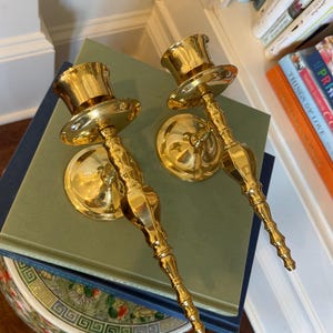 May include: Two gold-colored wall sconces with a classic design. Each sconce features a circular base, a decorative stem, and a top shaped like a small top hat. The sconces are made of a shiny, reflective material.