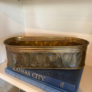 May include: A rectangular brass planter with an oval shape, featuring a decorative pattern of teardrop shapes. The planter is placed on top of two blue books, one of which reads "Kansas City". The planter is a vintage item.