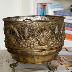 May include: A round brass planter with a textured surface featuring embossed shell and berry motifs. The vessel has a wide rim, a smooth lower section, and stands on three small, claw-shaped feet. The metal shows a weathered, aged patina.