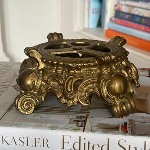 May include: A gold-toned metal stand featuring ornate, scroll-like detailing and four claw-style feet. The top surface has a circular cutout design with three smaller circular openings. The stand has a weathered, metallic finish with deep grooves.