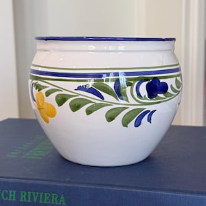 May include: A white ceramic bowl with a blue rim and floral design. The bowl features yellow and blue flowers with green leaves. The bowl is sitting on a blue book.