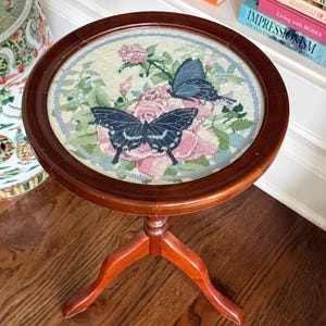 Op de afbeelding: Ronde, houten bijzettafel met een naaldkunst bovenblad. Het naaldkunstontwerp toont roze rozen en twee vlinders met zwarte en blauwe vleugels. De tafel heeft een donkerbruine afwerking en drie gebogen poten.