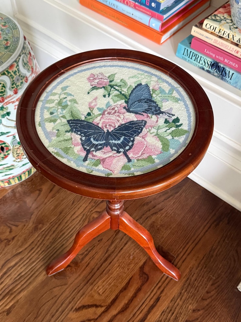 Op de afbeelding: Ronde, houten bijzettafel met een naaldkunst bovenblad. Het naaldkunstontwerp toont roze rozen en twee vlinders met zwarte en blauwe vleugels. De tafel heeft een donkerbruine afwerking en drie gebogen poten.