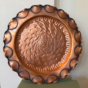 May include: A round copper tray with a scalloped edge and a textured, swirling pattern in the center. The rim features a decorative rope-like design. The tray's color is a warm copper with darker accents.
