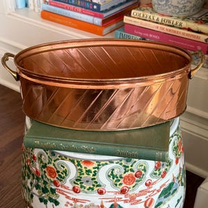 May include: A copper-colored, oval-shaped planter with diagonal etched lines and brass handles. The planter sits atop a decorative ceramic garden stool with a colorful floral and landscape design. Books are visible in the background.