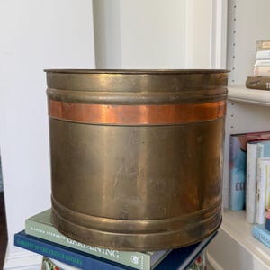 May include: A cylindrical brass planter with a copper band. The planter is sitting on a stack of books, including "Gardens of the French Riviera." The planter has a vintage look and is suitable for indoor or outdoor use.