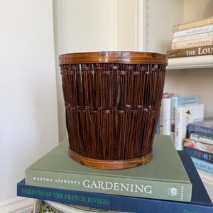 May include: A woven brown bamboo basket with a dark brown rim. The basket is sitting on top of two books, one green and one blue, with the text "GARDENING" and "GARDENS OF THE FRENCH RIVIERA" visible. The basket is on a decorative ceramic stool.