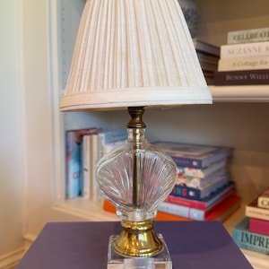 May include: A decorative table lamp with a pleated cream-colored lampshade. The lamp features a clear, textured glass body with a gold-colored metal base and accents. The lamp sits on a clear, square base. The lamp is approximately 18 inches tall.