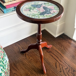 Op de afbeelding: Kleine, ronde, houten bijzettafel met een donkerbruine afwerking. Het tafelblad heeft een borduurontwerp van vlinders en bloemen in blauwe, roze en groene tinten. De tafel heeft een enkele centrale steun en drie poten.
