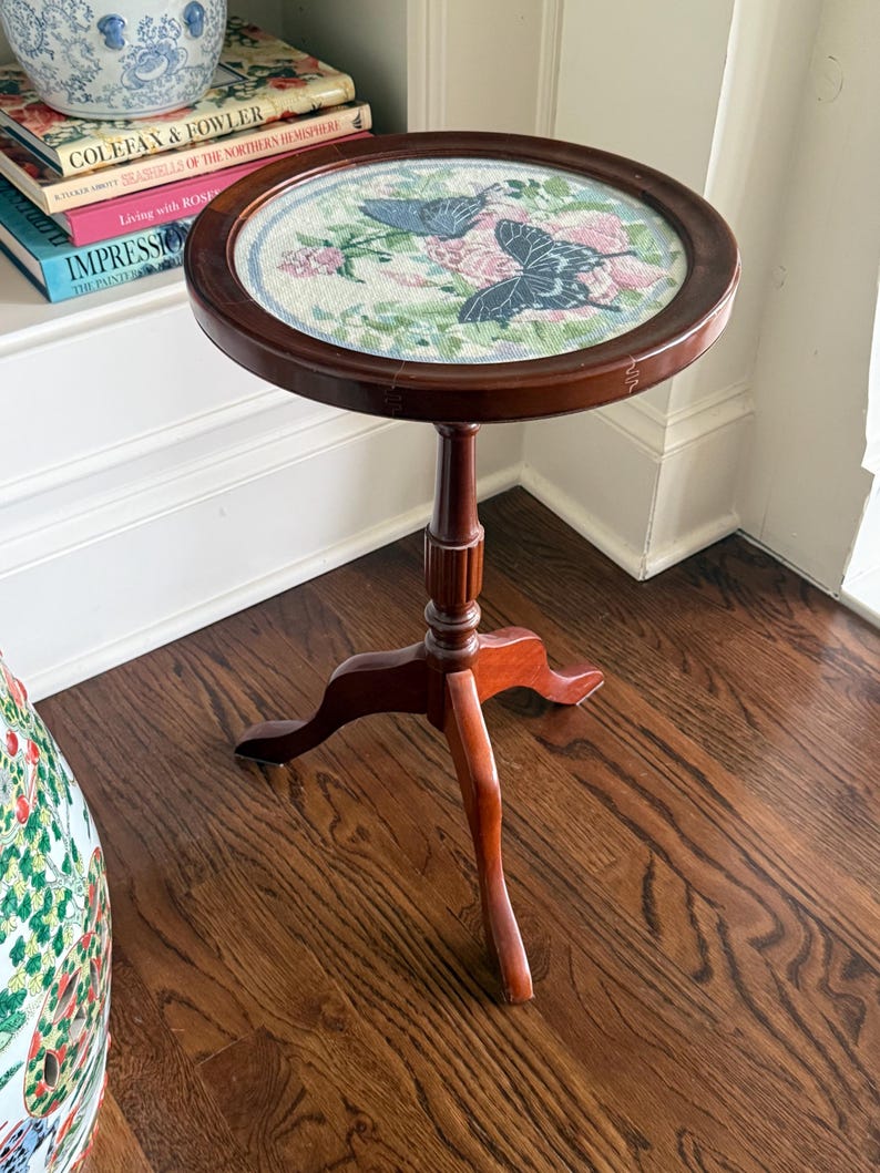 Op de afbeelding: Kleine, ronde, houten bijzettafel met een donkerbruine afwerking. Het tafelblad heeft een borduurontwerp van vlinders en bloemen in blauwe, roze en groene tinten. De tafel heeft een enkele centrale steun en drie poten.