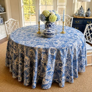 May include: A round dining table covered in a blue and white floral patterned tablecloth. Centered on the table is a blue and white ceramic vase filled with green and blue hydrangeas, a lidded ceramic tureen, and two tall brass candle holders.