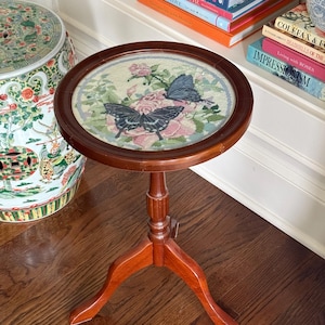 Op de afbeelding: Een kleine, ronde houten bijzettafel met een donkerbruine afwerking. Het tafelblad heeft een borduurontwerp van roze rozen en twee vlinders. De tafel heeft een driepoot en is ongeveer 61 cm hoog.