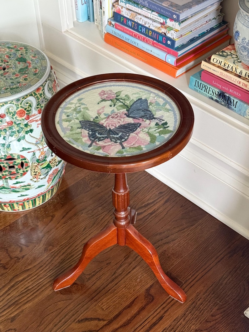 Op de afbeelding: Een kleine, ronde houten bijzettafel met een donkerbruine afwerking. Het tafelblad heeft een borduurontwerp van roze rozen en twee vlinders. De tafel heeft een driepoot en is ongeveer 61 cm hoog.