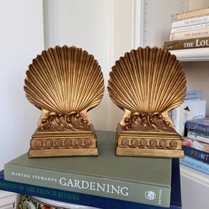 May include: Two gold-colored seashell bookends. Each bookend features a detailed shell design with a wave and decorative base. The bookends are displayed on top of a green and blue book.
