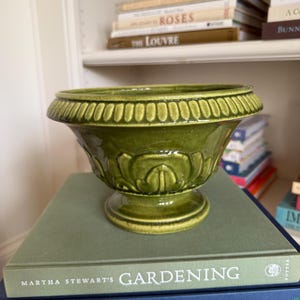 May include: A green ceramic planter with a pedestal base and decorative rim. The planter is sitting on a stack of books, including one titled "MARTHA STEWART'S GARDENING". The planter has a detailed floral design.