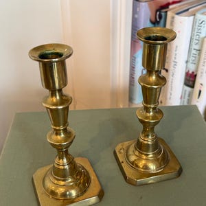 Puede incluir: Dos candelabros antiguos de lat&oacute;n. Cada candelabro tiene una base cuadrada, un tallo decorativo y una parte superior circular para sostener una vela. El lat&oacute;n tiene un tono dorado c&aacute;lido y el dise&ntilde;o es cl&aacute;sico y elegante.