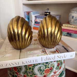 May include: Two matching brass bookends shaped like large, ribbed seashells. Each piece features a metallic gold finish with vertical ridges and a rounded, fan-like silhouette. The bookends are positioned on top of a white hardcover book titled Suzanne Kasler Edited Style.