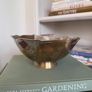 May include: A decorative, antique-style bowl with a scalloped rim and a gold-colored base. The bowl has a tarnished, metallic finish. It sits atop a stack of books, including one titled "Gardening."