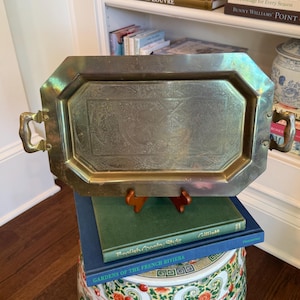 May include: A vintage brass tray with ornate detailing and handles. The tray is rectangular with an octagonal border. It rests on a stack of books, including "English Country Style" and "Gardens of the French Riviera."
