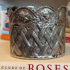 May include: A silver-colored, decorative container with a woven basket design on the body and a floral pattern around the scalloped rim. The container sits on a book titled "Glory of Roses".