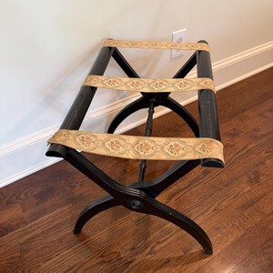 May include: A black luggage rack with decorative floral patterned straps. The rack has a classic design with curved legs and a central support. The straps are beige with a repeating pattern of flowers and leaves. The rack is made of wood.