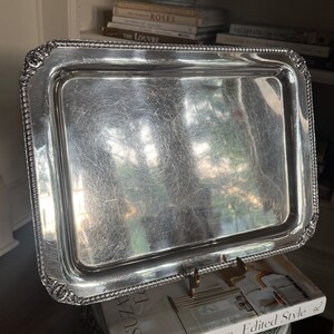 May include: A rectangular silver-toned serving tray with a polished, reflective surface and a decorative beaded border. The edges feature ornate, embossed floral detailing at each corner, providing a textured contrast to the smooth center.