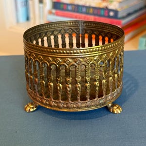 May include: Ornate, antique brass-colored metal container with a circular design. The container has a decorative openwork pattern around the sides and rests on three small, claw-shaped feet. The interior is visible, and the piece has a vintage aesthetic.