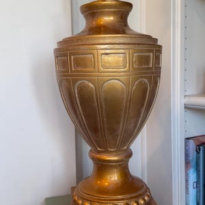 May include: A gold-colored table lamp with a classic urn shape. The lamp features a square base, a detailed body with rectangular and arched designs, and a brass-colored lamp holder. The lamp is approximately 24 inches tall.