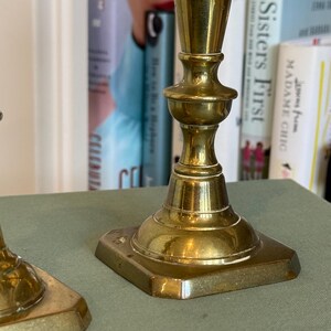 Puede incluir: Dos candelabros antiguos de lat&oacute;n con base cuadrada y detalles ornamentados. Los candelabros tienen un acabado pulido y se exhiben sobre una superficie verde. El fondo incluye una estanter&iacute;a con libros.