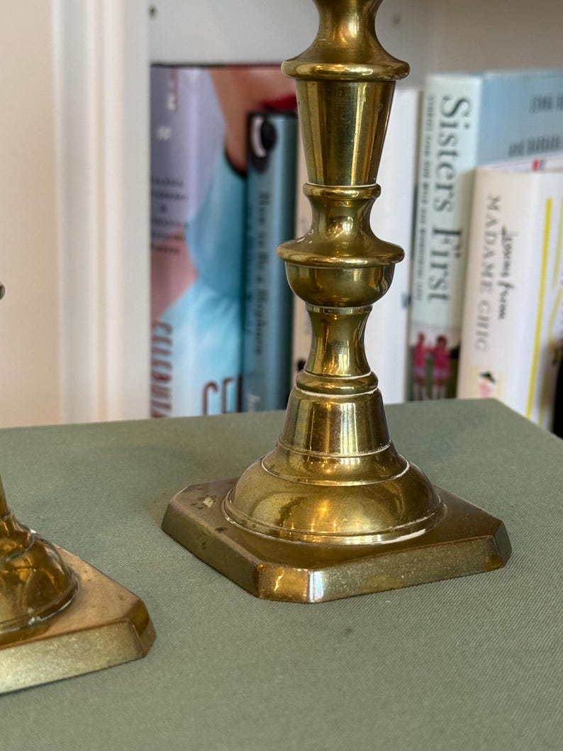 Puede incluir: Dos candelabros antiguos de lat&oacute;n con base cuadrada y detalles ornamentados. Los candelabros tienen un acabado pulido y se exhiben sobre una superficie verde. El fondo incluye una estanter&iacute;a con libros.