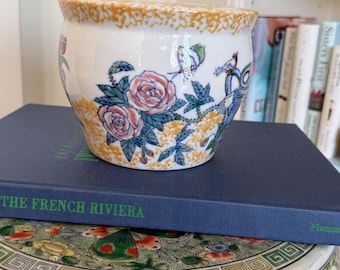 Puede incluir: Una maceta de cerámica blanca con un diseño floral con rosas rosas, hojas azules y pájaros. El borde tiene un patrón texturizado, moteado de color naranja-amarillo. La maceta está sobre un libro azul titulado "THE FRENCH RIVIERA".