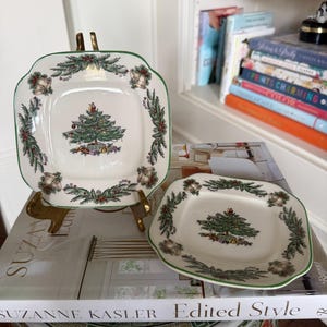 May include: Two square ceramic plates featuring a central Christmas tree illustration surrounded by a border of green pine branches, red berries, and gold bells. The plates have a cream-colored base with a thin green rim along the edges.