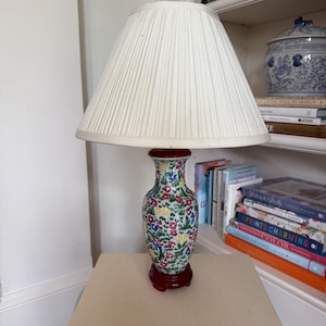 May include: A decorative table lamp with a white pleated lampshade. The lamp base is a colorful floral vase with a dark red base. The lamp is sitting on a book titled "The Glory of Roses".